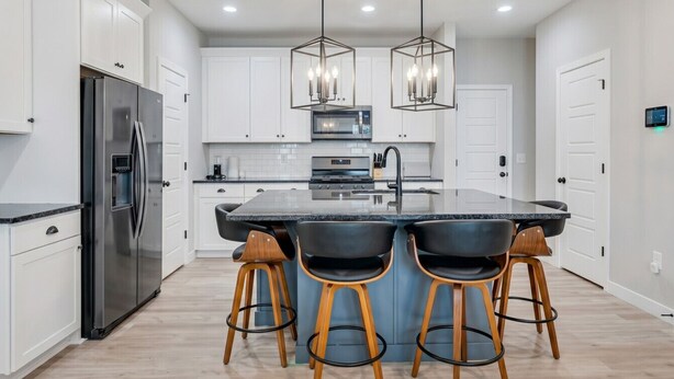 A modern kitchen with white cabinets, a stainless steel refrigerator, a central island with bar stools, and pendant lighting.