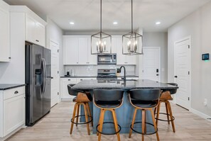 A modern kitchen with white cabinets, a stainless steel refrigerator, a central island with bar stools, and pendant lighting.