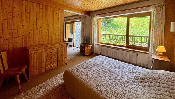 Een houten kamer met een bed, een bureau en een raam met uitzicht op groen.