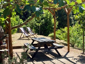 Eine hölzerne Pergola mit Picknicktisch und zwei Liegestühlen, umgeben von Grünpflanzen.