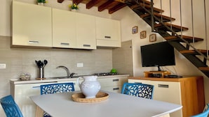 A kitchen with white cabinets, a dining table with blue chairs, a TV, and a wooden staircase. - Three-room 2-bedroom apartment and sea view terrace (Rio Nell'elba)