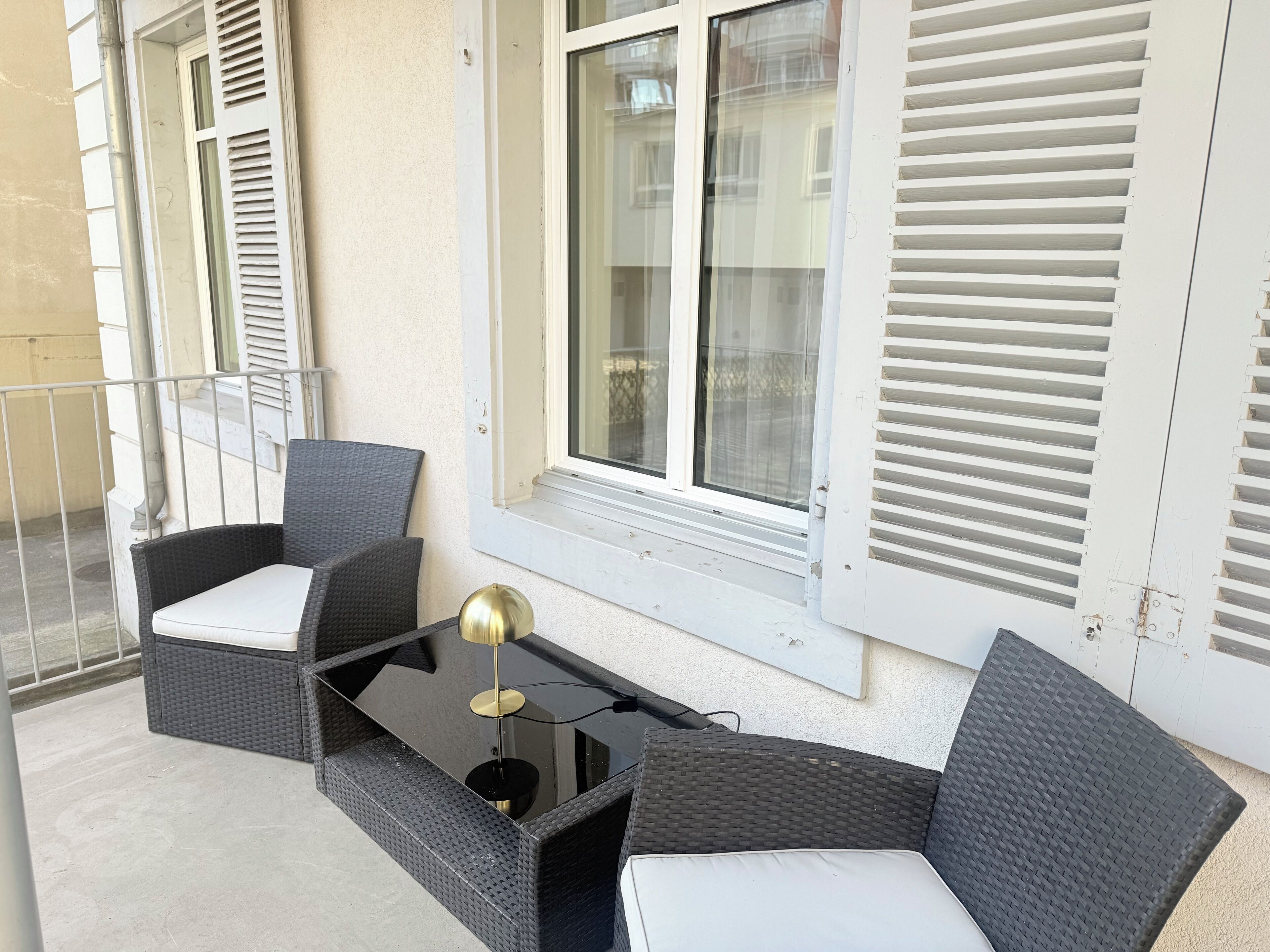 A balcony with wicker furniture, a glass table, and a window with shutters.