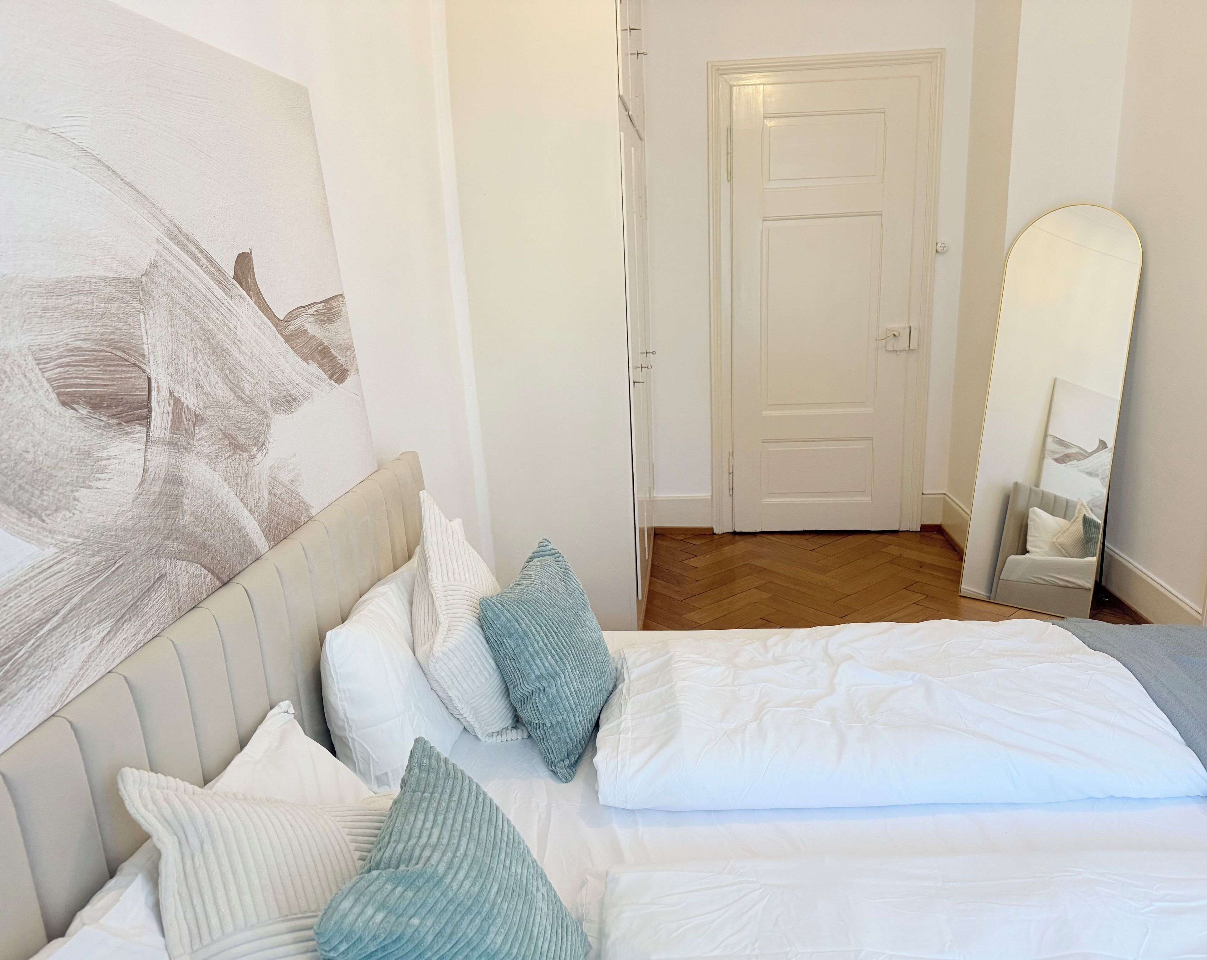 A bedroom with a bed, pillows, a painting, a door, and a mirror.