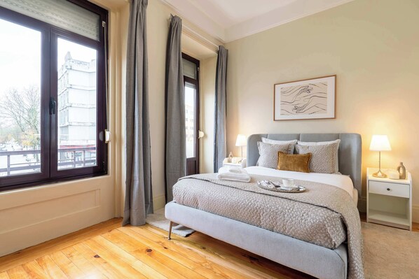 Ein Schlafzimmer mit Bett, Nachttischlampen, einem Fenster mit Vorhängen, einem Heizkörper und einem gerahmten Bild an der Wand.