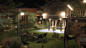 Een houten tuinhuisje met een bordje in een vreemde taal, omgeven door een goed verlichte tuin met tafels en stoelen.