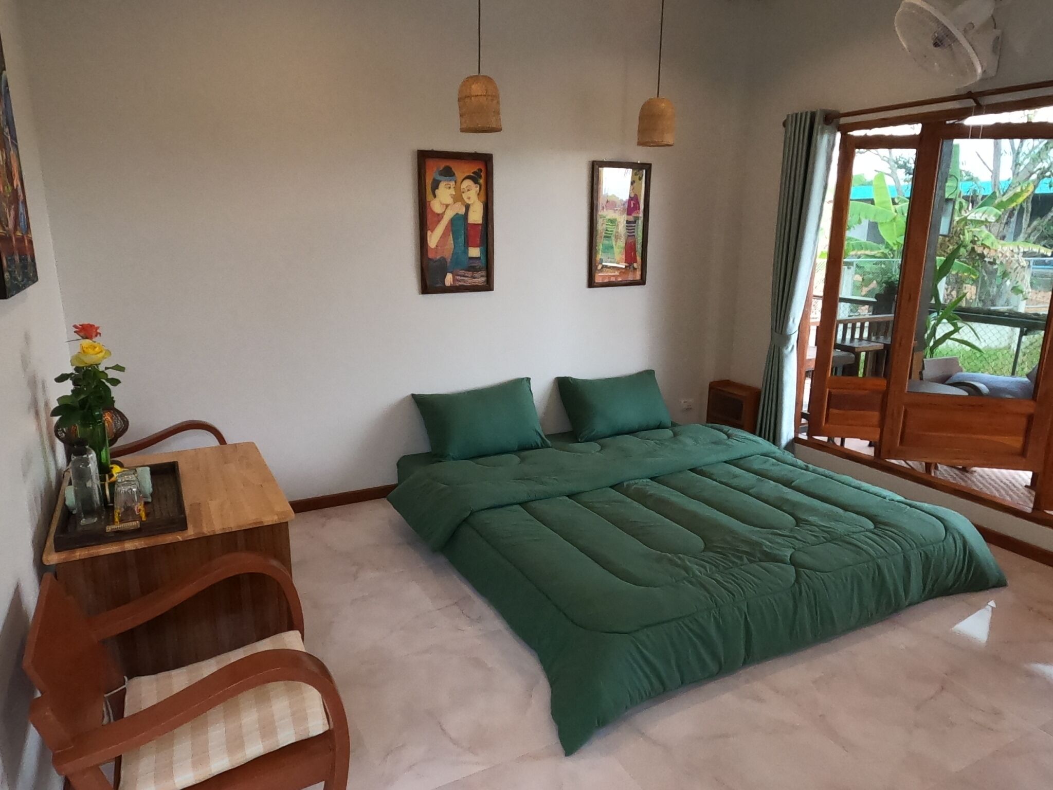 Ein Schlafzimmer mit einem großen Bett, einem hölzernen Schreibtisch mit Blumen in einer Vase, zwei an der Wand hängenden Bildern und Blick ins Freie durch eine Schiebetür.