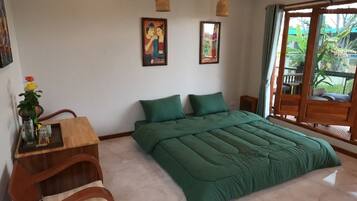 Ein Schlafzimmer mit einem großen Bett, einem hölzernen Schreibtisch mit Blumen in einer Vase, zwei an der Wand hängenden Bildern und Blick ins Freie durch eine Schiebetür.