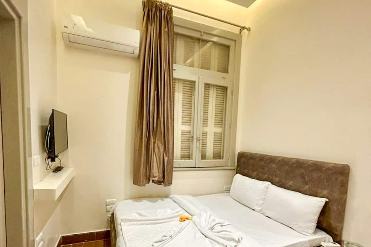 A bedroom with a bed, a TV on a shelf, a window with curtains, and a thermostat on the wall.