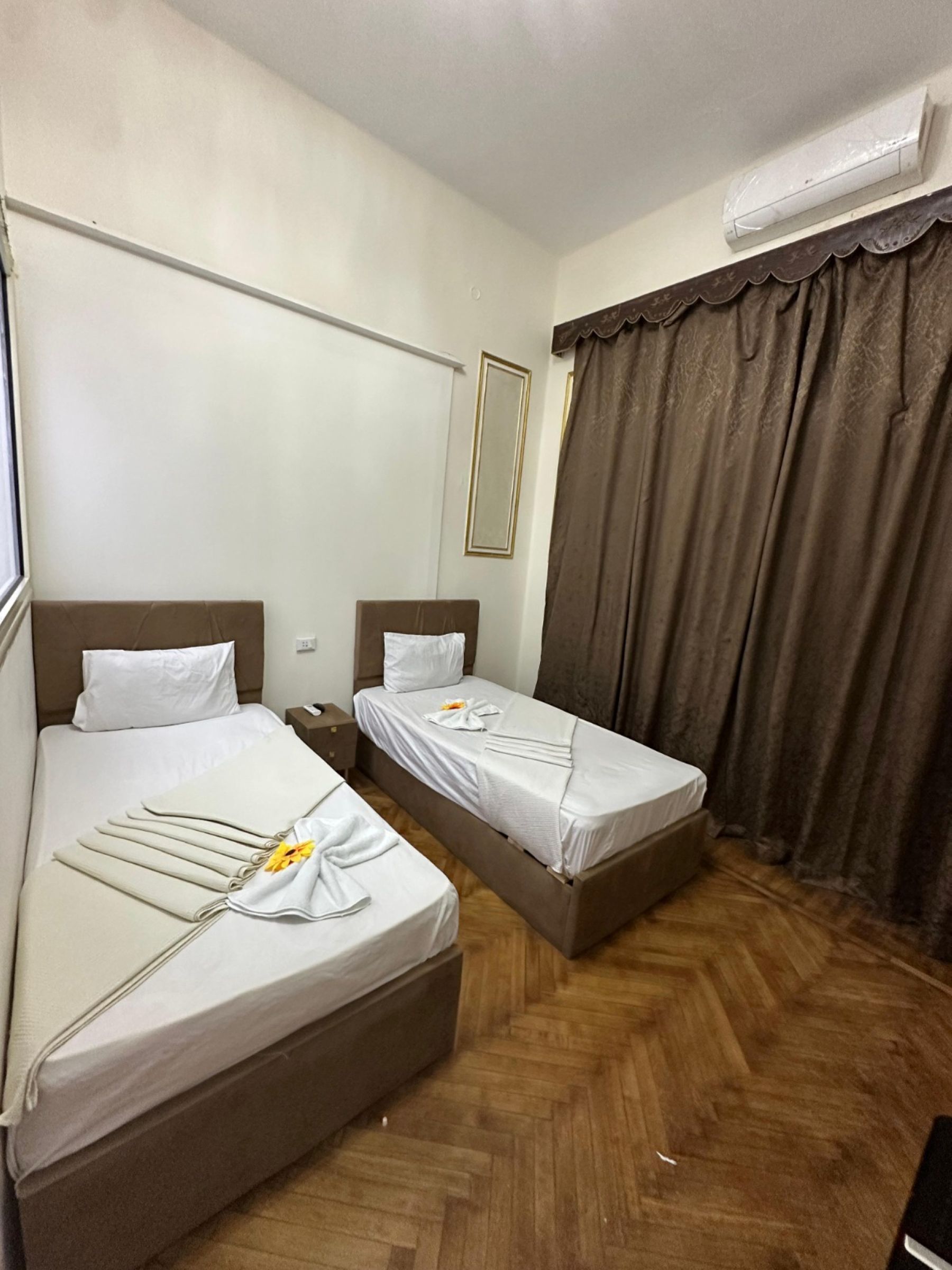 A hotel room with two beds, a mirror, an air conditioning unit, and a window with curtains.