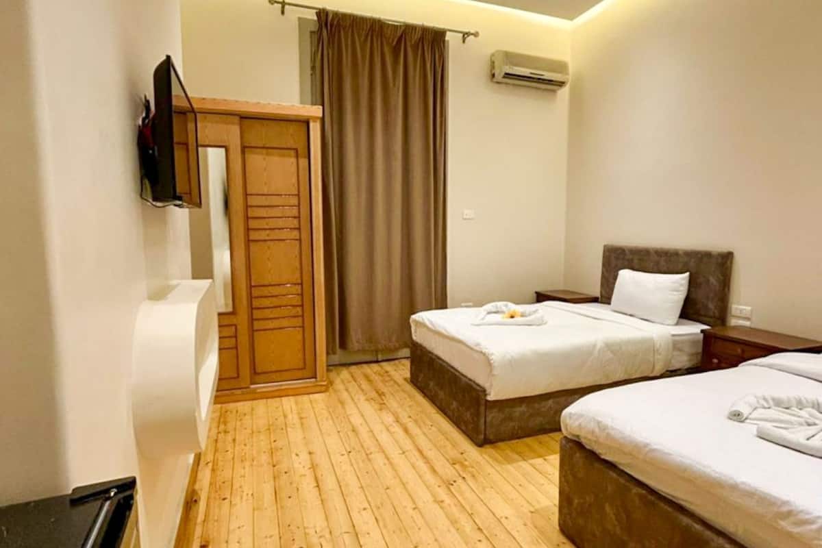 A hotel room with two beds, wooden flooring, a TV mounted on the wall, an air conditioning unit, and a wooden wardrobe.