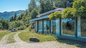 Una casa moderna con amplios ventanales de vidrio, un camino de adoquines y montañas al fondo.