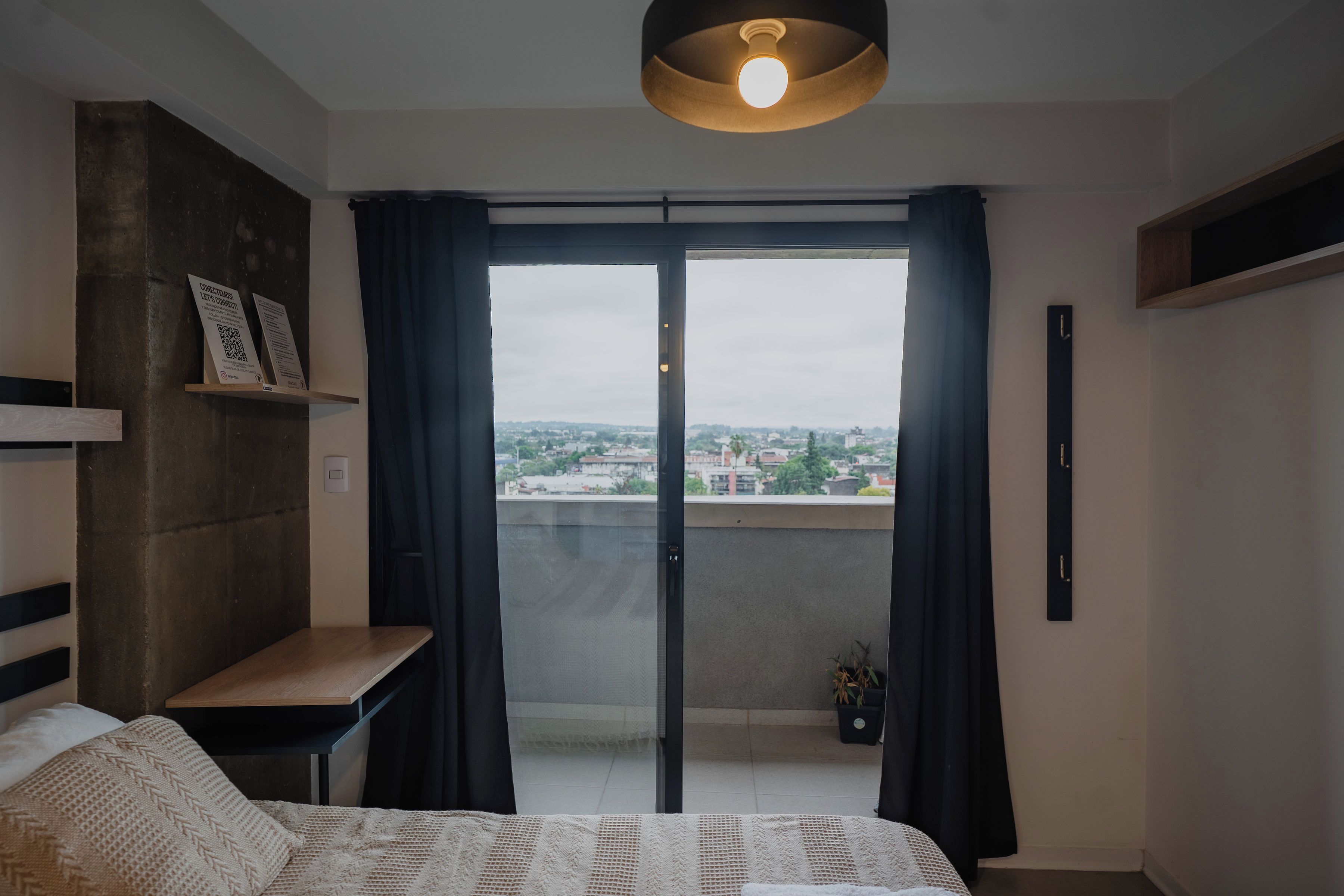 Une chambre moderne avec un lit, une petite table, un luminaire suspendu et une vue sur le paysage urbain, visible à travers une porte vitrée.