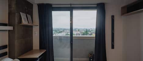 Quarto moderno com cama, uma mesinha, luminária pendente e vista para o horizonte urbano através de uma porta de vidro.