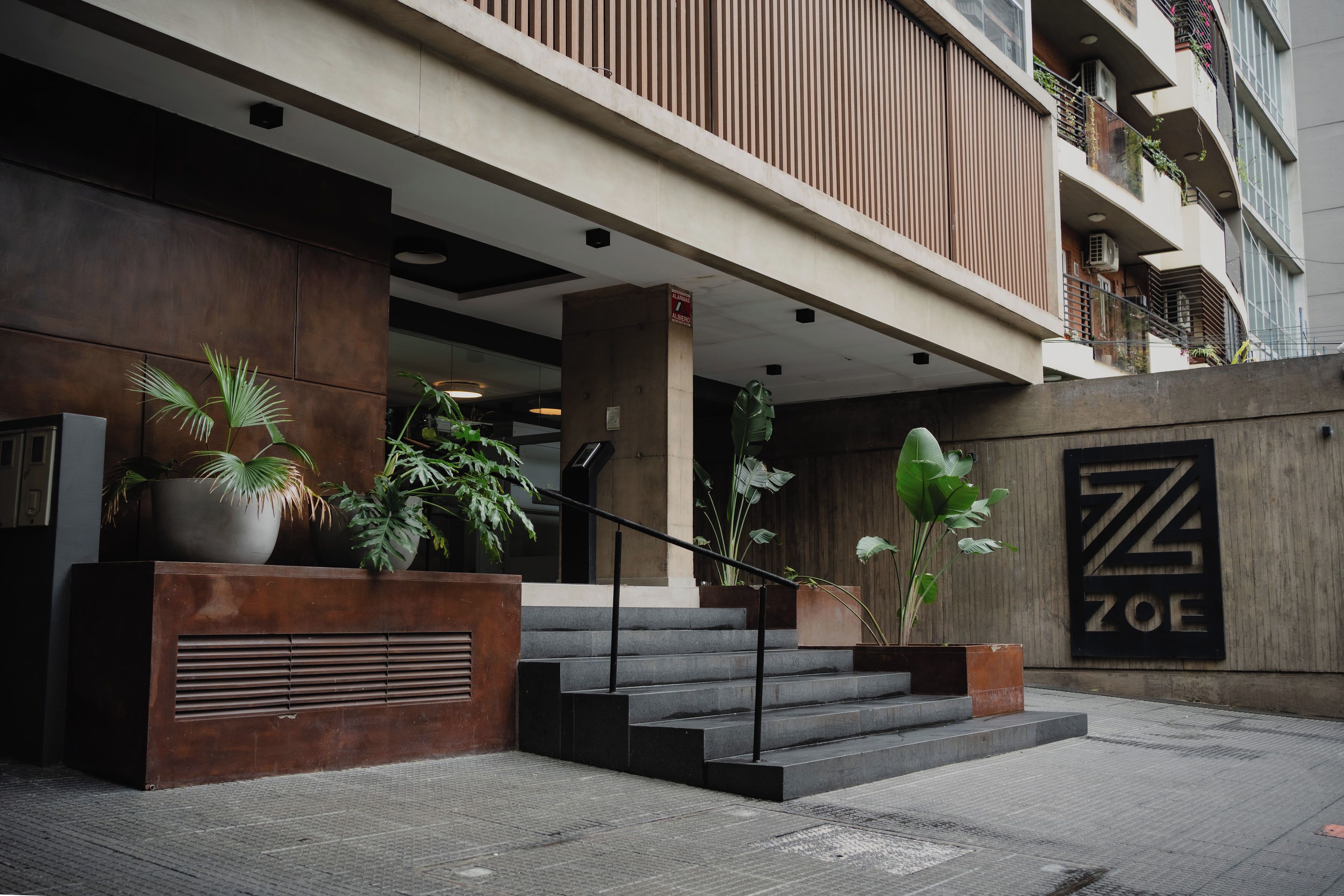 Entrada de hotel con diseño moderno, que incluye una escalera hacia la entrada, plantas en macetas y un cartel con el nombre del hotel.