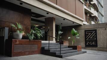 Entrada de hotel con diseño moderno, que incluye una escalera hacia la entrada, plantas en macetas y un cartel con el nombre del hotel.