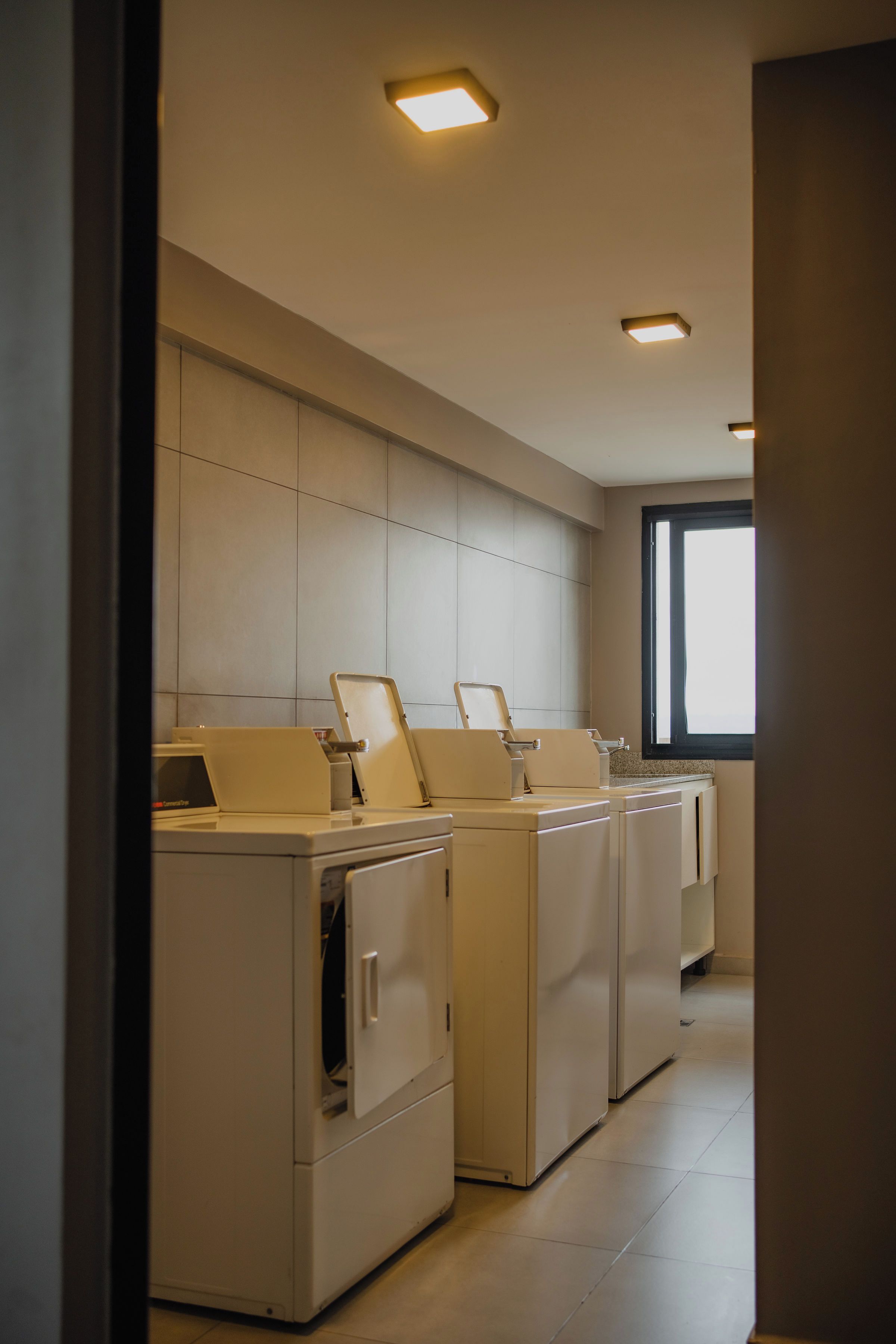 Un cuarto de lavandería con varias lavadoras y secadoras, piso de baldosas y una ventana que proporciona luz natural.