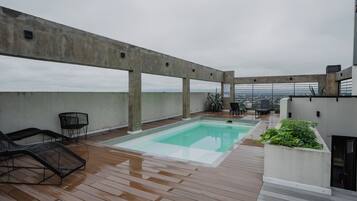 Un área de piscina en la azotea con terraza de madera, sillones de descanso y vistas a la ciudad.