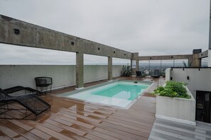 Un área de piscina en la azotea con terraza de madera, sillones de descanso y vistas a la ciudad.