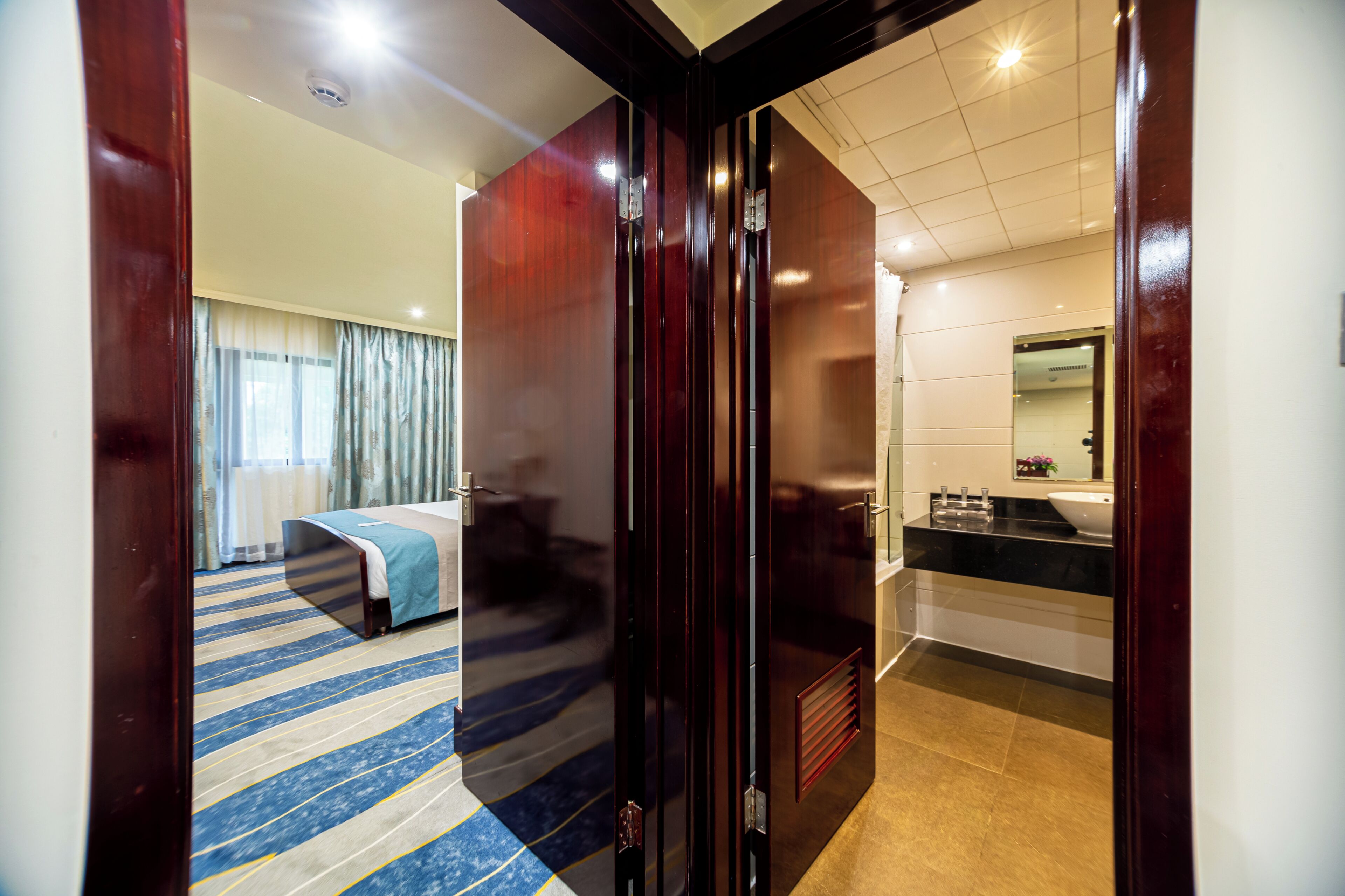 Una habitación de hotel con cama, baño con lavamanos y espejo, y piso alfombrado.