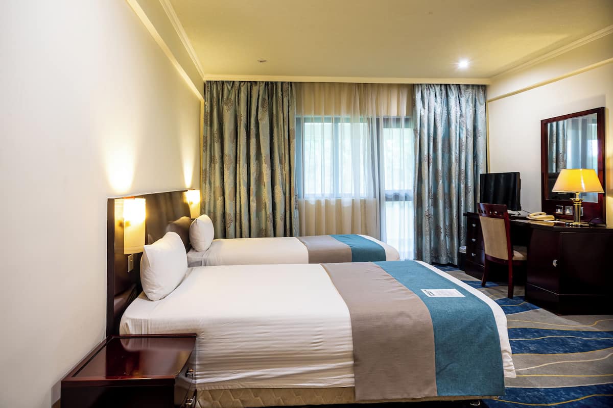 A hotel room with two beds, a desk, a chair, a lamp, and a large window with curtains.
