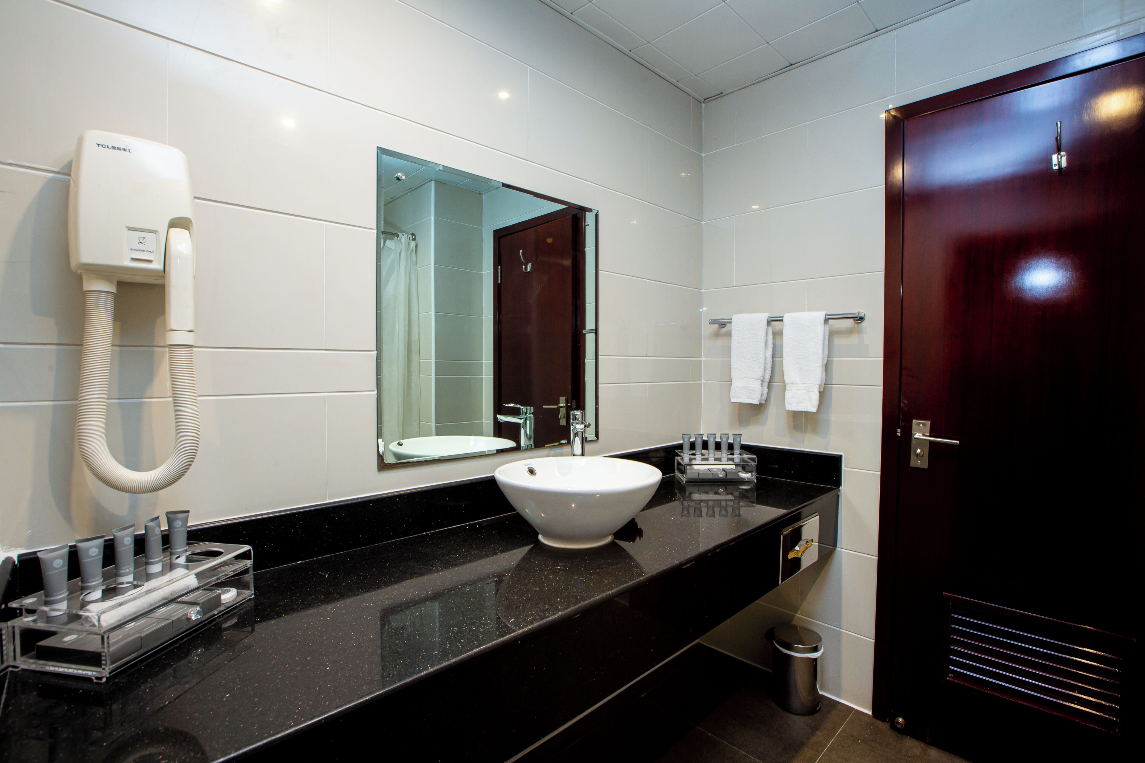 A modern bathroom with a black countertop, a white sink, a handheld showerhead, a mirror, and a dark wooden door.