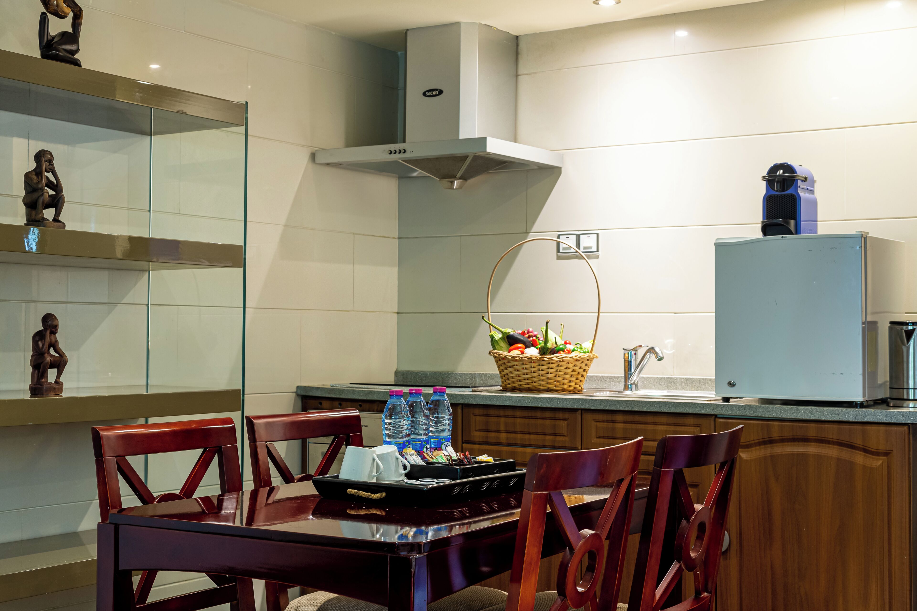 A modern kitchen with a dining area, featuring a stainless steel range hood, a refrigerator, and a basket of fresh produce.