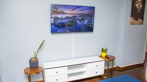 Et værelse med et fladskærms-TV monteret på væggen, et hvidt konsolbord med skuffer, to små sengeborde med potteplanter og en blå tæppe på gulvet.