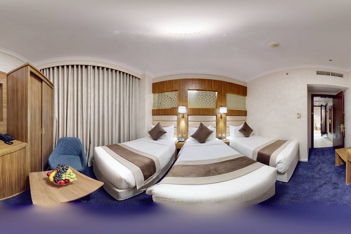 A hotel room with a curved wooden desk, a flat-screen TV, two beds with headboards, a blue armchair, and a fruit bowl.