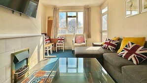 A living room with a fireplace, a sofa with British flags, a flat-screen TV, and a window with curtains. - Spacious Flat in Nine Elms (London)