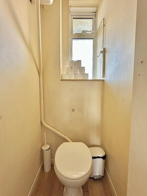 A small, narrow bathroom with a toilet, a trash bin, and a window. - Spacious Flat in Nine Elms (London)
