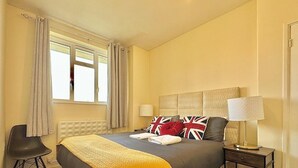 A hotel room with a large bed, a bedside table with a lamp, a chair, and a window with curtains. - Spacious Flat in Nine Elms (London)
