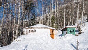 Un paysage enneigé avec une petite construction en bois, peut-être une cabane, entourée d’arbres et sous un ciel dégagé.