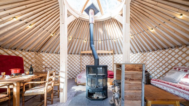 L’intérieur d’une yourte, avec un poêle à bois central, une table à manger, des chaises et un lit.