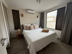 A bedroom with a large bed, an air conditioning unit, a window with curtains, and a view of the outside.