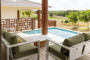 Zona junto a la piscina con dos sillones verdes acolchados, una mesa blanca y vista a un jardín ajardinado.