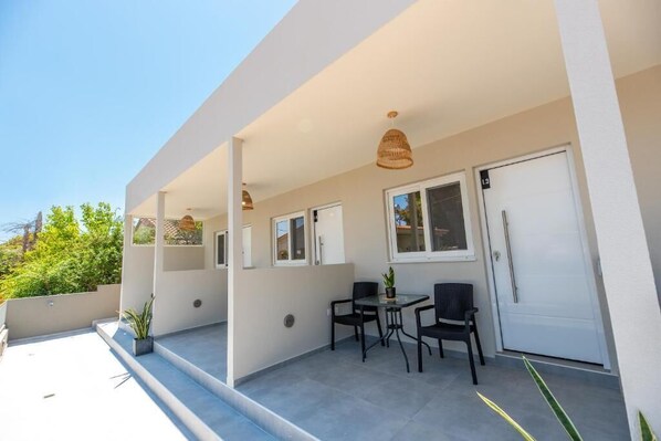 A modern outdoor patio with a glass table and chairs, a white door, and a hanging light fixture. - AMELI COZY APARTMENTS  (Spata-Artemida)