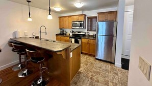 A modern kitchen with a central island, bar stools, stainless steel appliances, wooden cabinets, and pendant lighting. - Aspens Retreat - 1BR w/Hot Tub (Golden)