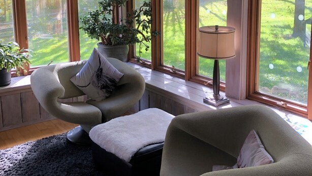 Een gezellige kamer met een gebogen bank, een klein tafeltje en een potplant. Grote ramen bieden uitzicht op een groene tuin.