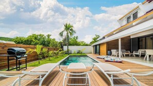 Patio trasero con piscina, dos sillones, una parrilla y un área de patio con mesa y sillas.
