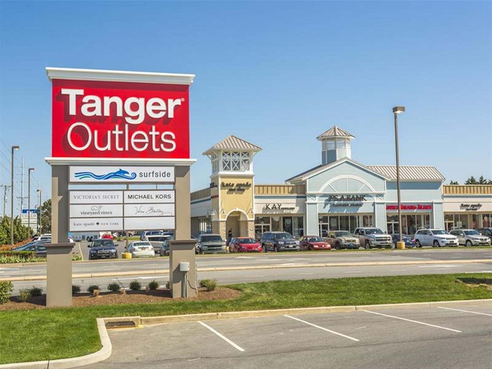 Ett köpcentrum som tillhör kedjan Tanger Outlets, med en stor skylt, en parkeringsplats och ett antal olika butiker.