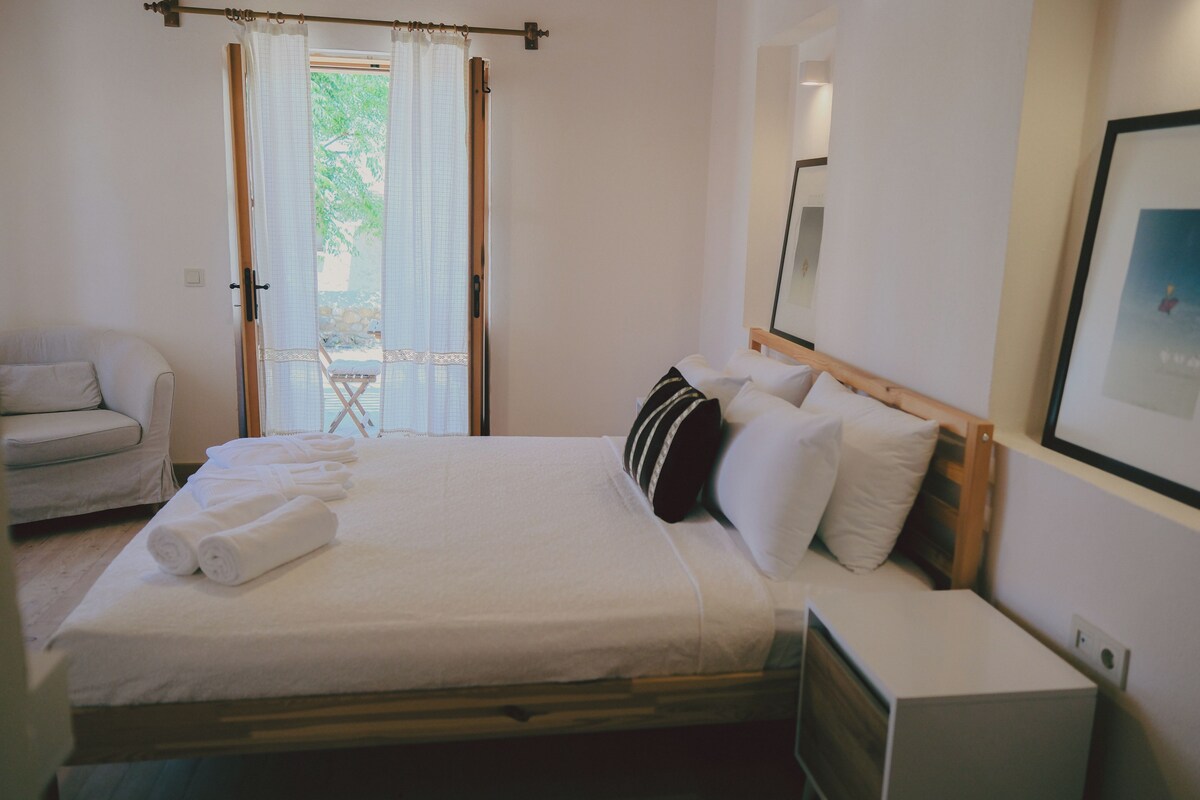 A neatly made bed with white linens and a pillow, a bedside table with a lamp, and a chair in a room with a large window and a door.
