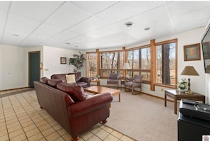A living room with a brown leather sofa, a coffee table, and a TV stand. There are large windows with a view of trees.