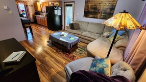A living room with a sofa, a coffee table, a lamp, and a dining area with a kitchen in the background.