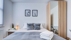 A bedroom with a bed, pillows, a bedside table with a lamp, a mirror, and two framed pictures on the wall. - Cornwall The Duchess 3 Bed Duplex 10 guests (London)