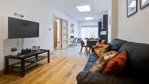 A modern living room with a flat-screen TV, a dark sofa with orange cushions, a wooden coffee table, and a dining area in the background. - Cornwall The Duchess 3 Bed Duplex 10 guests (London)
