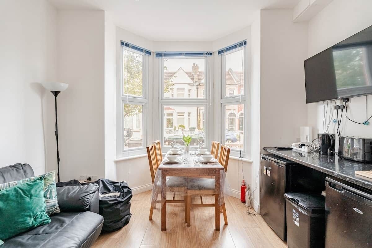 A compact living space with a dining table set for four, a sofa, a kitchen area with a TV, and large windows offering a view of the neighborhood.