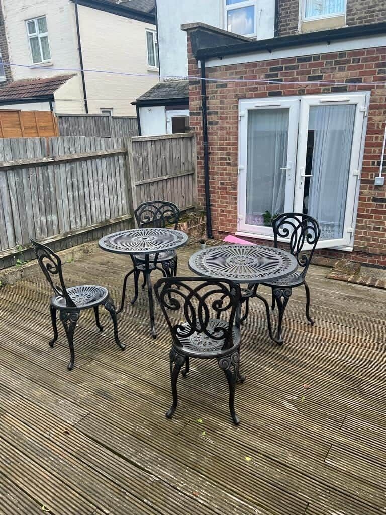 A patio with four metal tables and chairs, a wooden deck, a brick house, and a white-framed window.