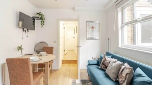 A modern living room with a blue sofa, a dining table with chairs, a TV mounted on the wall, and a door leading to another room. - Springfield - The Earl (London)