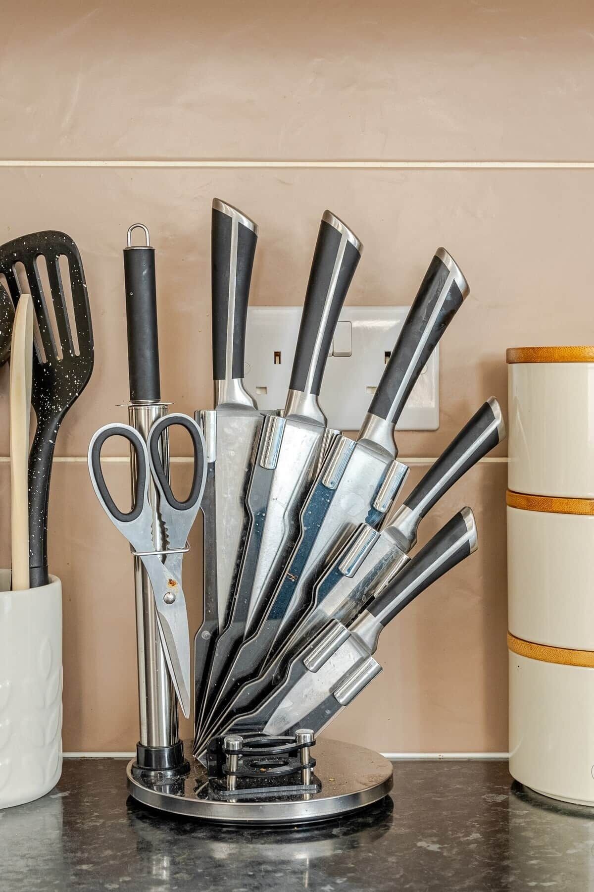 A kitchen utensil holder with various tools, a white container, and a power outlet on the wall.