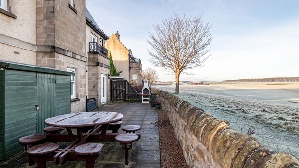 An outdoor patio with a wooden table and benches, a stone wall, and a view of a frozen body of water. - Golf Court (No 2) | Elie (Elie)